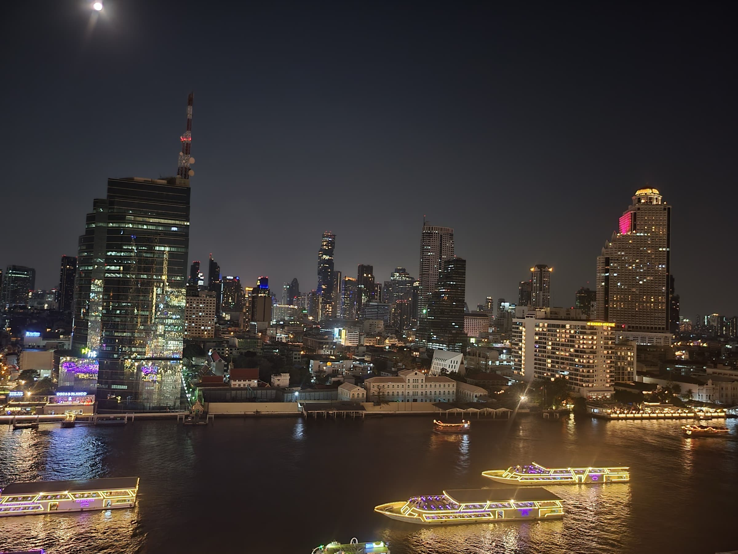 Bangkok's skyline at night across the Chao Phraya river, with lit river cruise boats